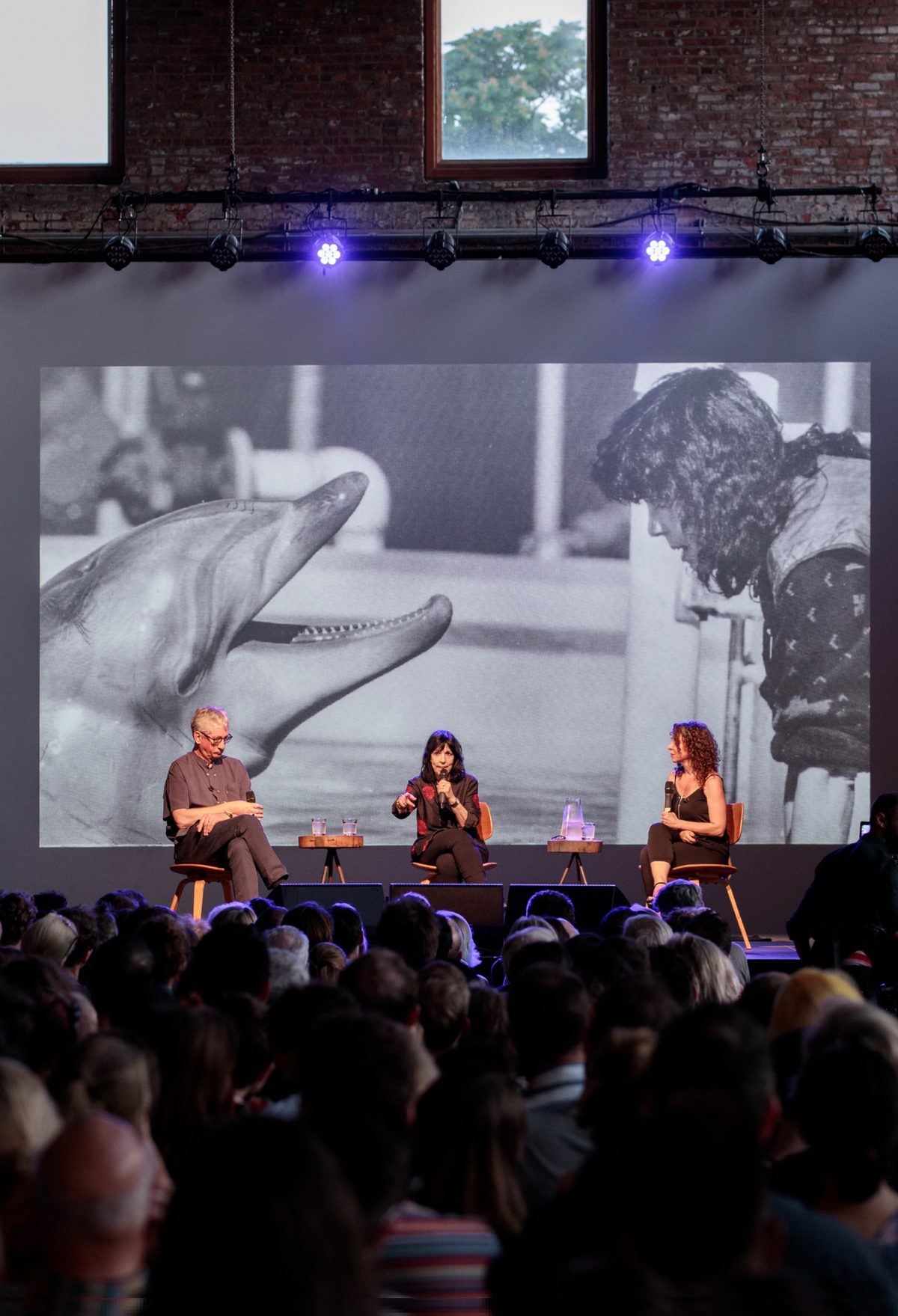 photo of large event space with speakers on the stage and a projection of a dolphin poking its head out of the water towards a trainer in the background